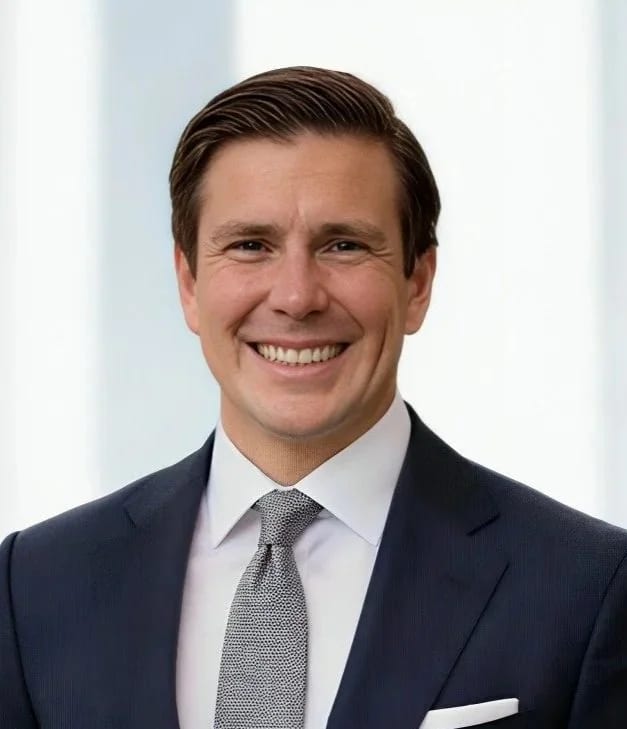 A smiling man in a dark suit, white dress shirt, and patterned tie, standing in a bright, modern office setting.