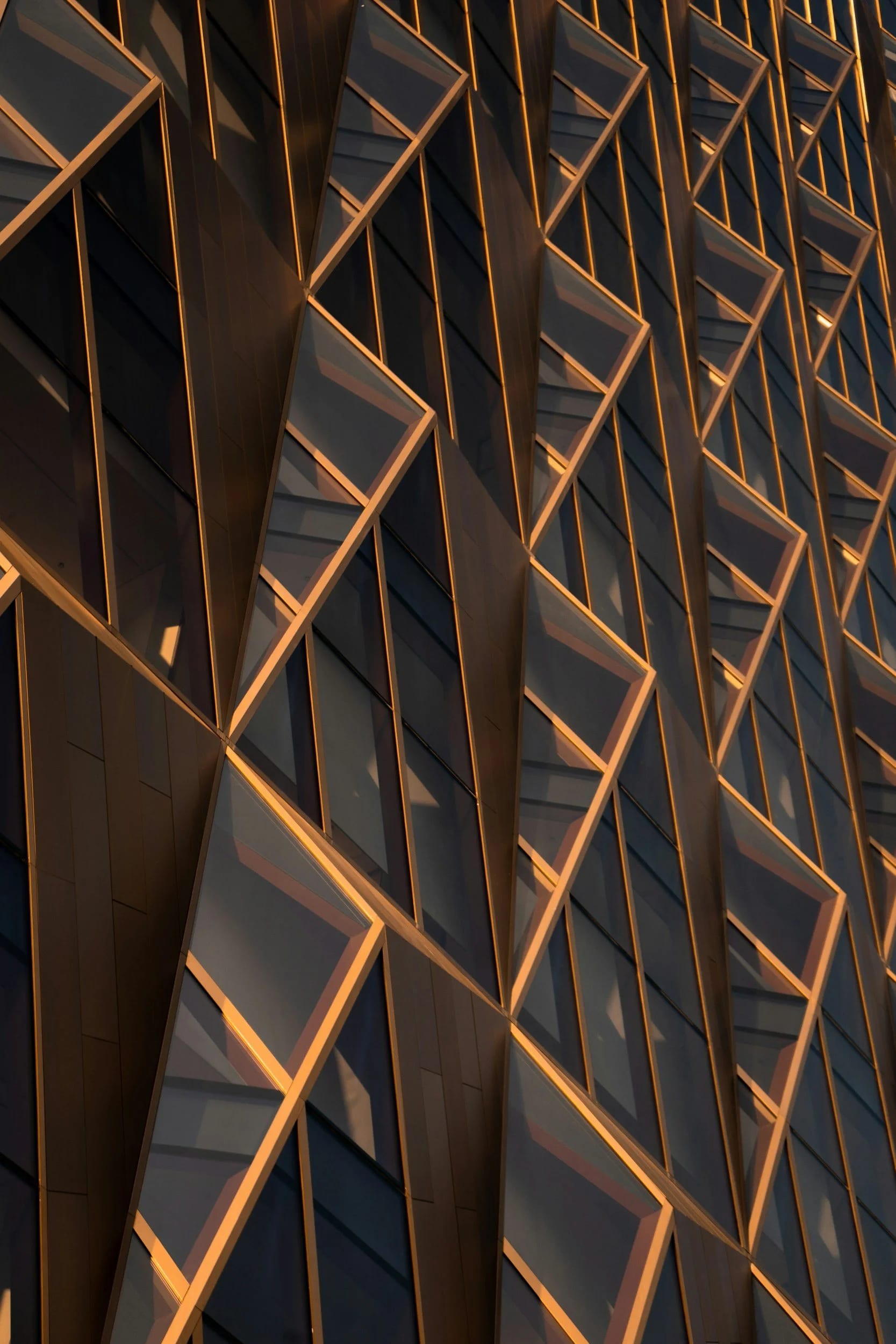 Close-up of a modern building's exterior featuring a geometric pattern of windows with metal frames, illuminated by warm lighting at dusk.