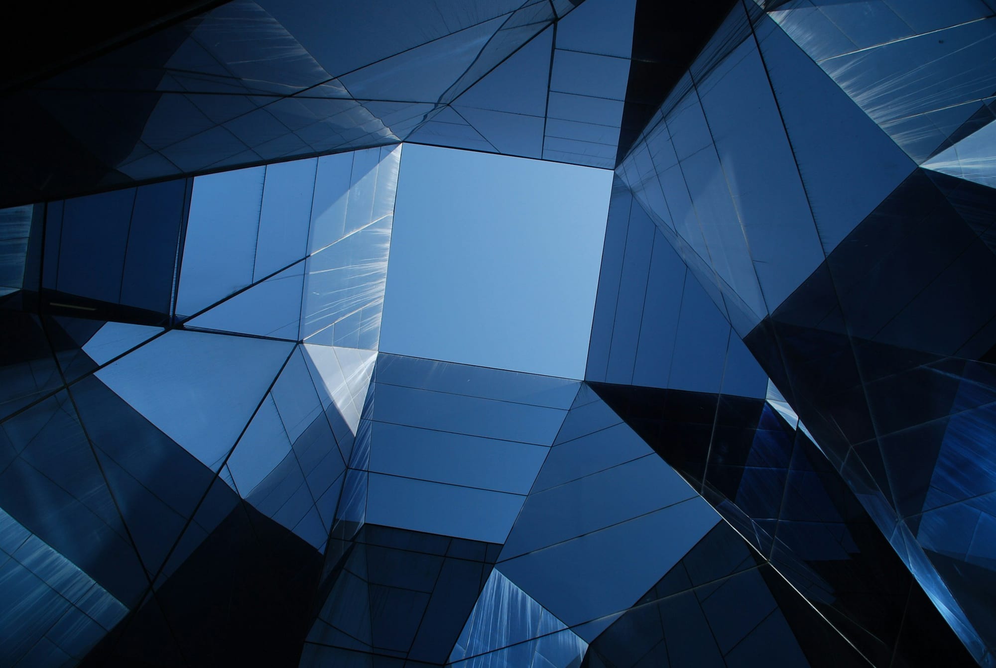 Looking up at the glass facade of a modern building with geometric shapes and reflections of the blue sky.