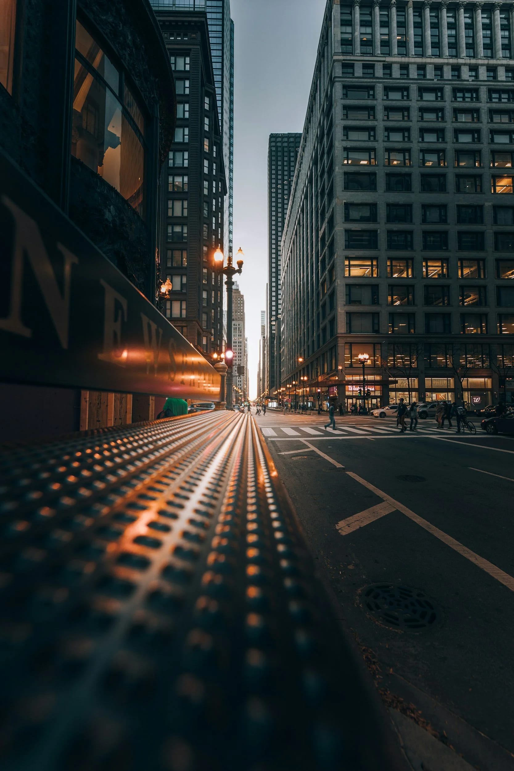 City street at sunset with tall buildings, pedestrians crossing the street, and streetlights glowing.